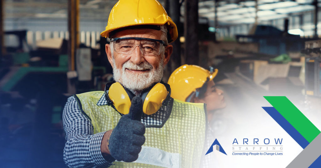 An older white male is wearing a yellow hard hat and giving a thumbs up